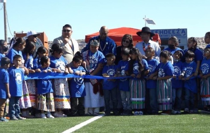 A large group of elementary school students and adults standing behind a blue ribbon that is being cut.